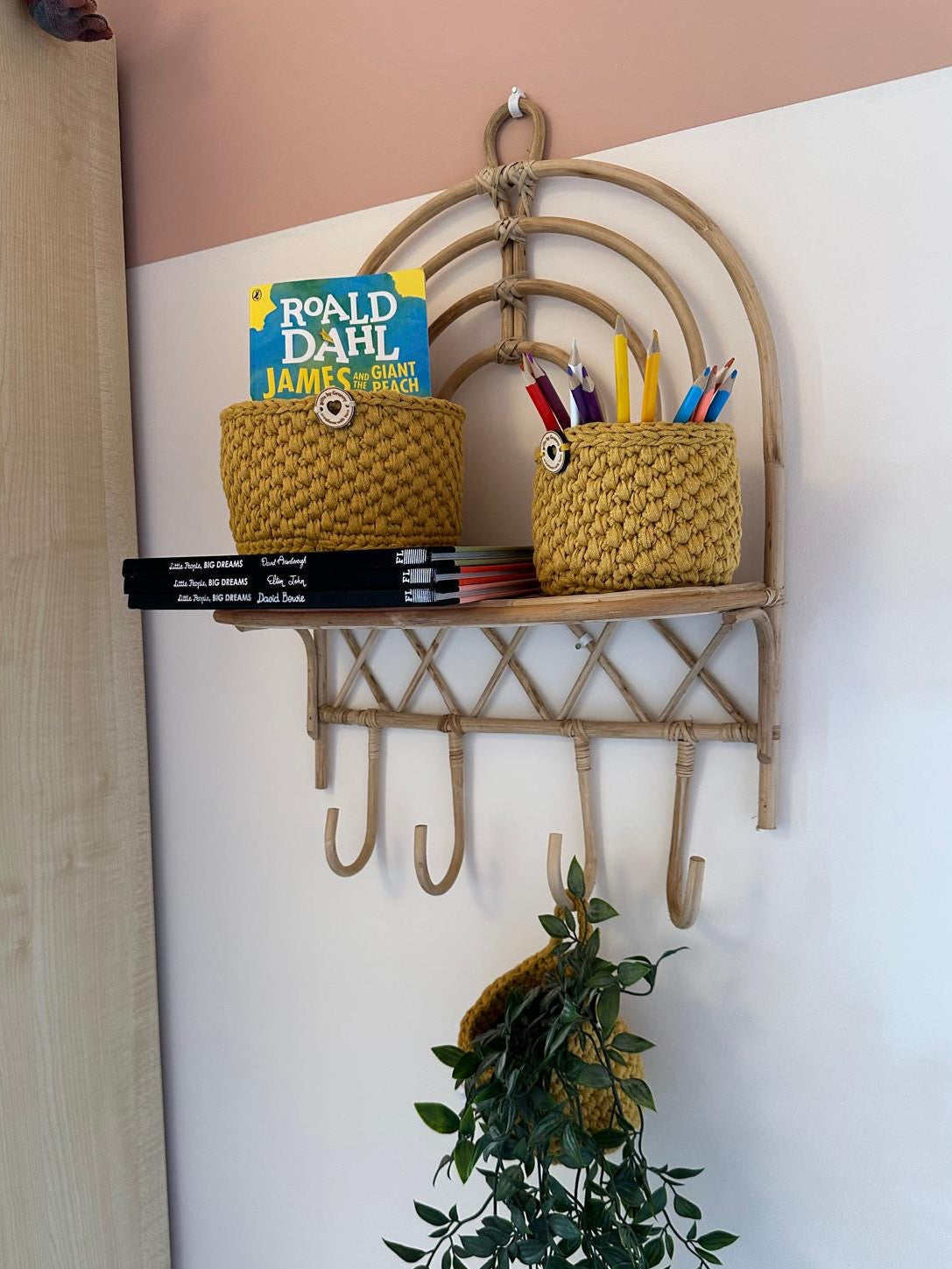 A natural bamboo wall-mounted shelf with five hooks, styled with two woven mustard-yellow baskets, books, and colorful pencils, set against a two-tone pink and white wall.