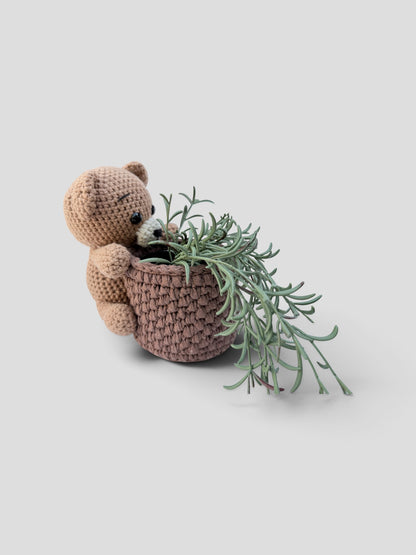 A crocheted teddy bear holding a small plant pot cover filled with green leafy stems, photographed on a plain light gray background.
