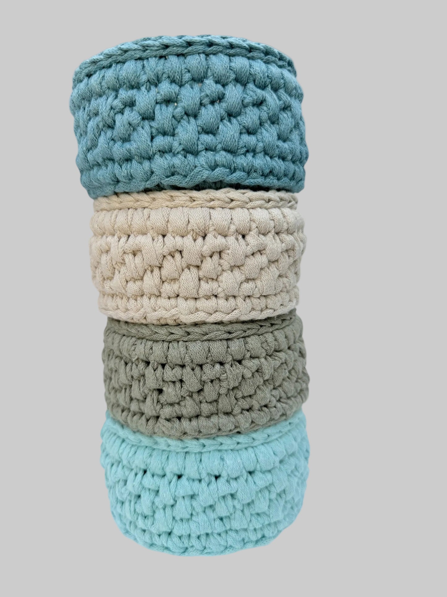 Four crocheted cylindrical storage baskets stacked vertically, featuring textured chunky crochet patterns in petrol blue, ecru, sage green and mint on a chalky white background.