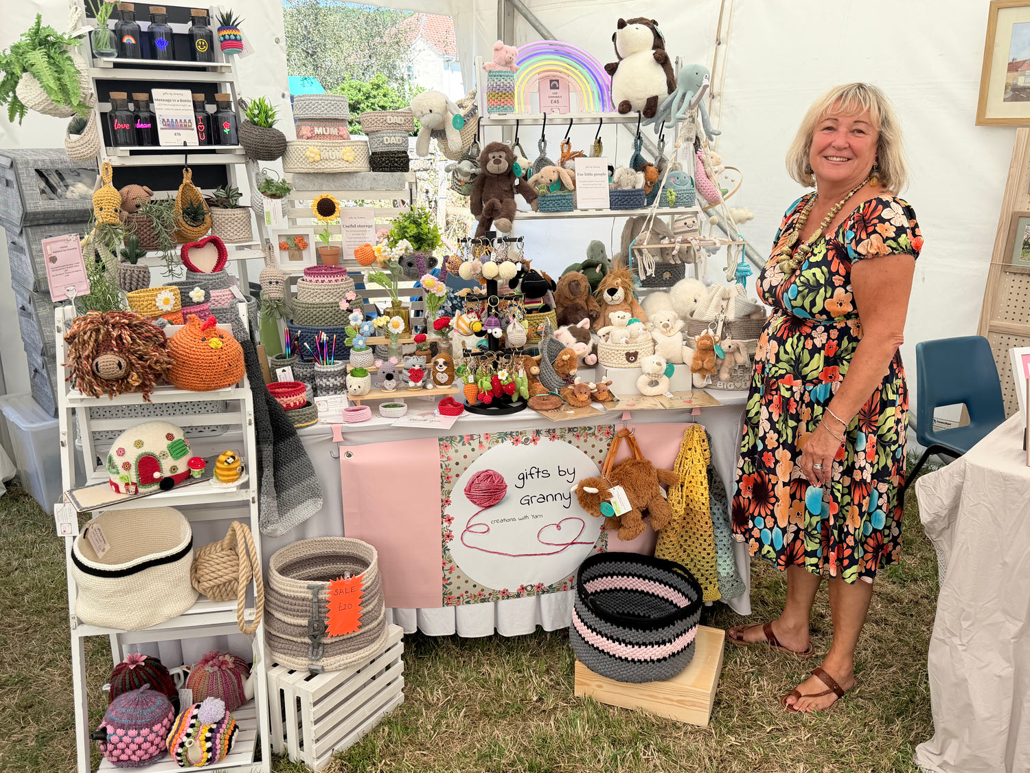 A woman stands beside a small market stall filled with handmade baby items, plush toys, baskets, and accessories under a white tent. The stall has a sign reading 'gifts by cecelia'.
