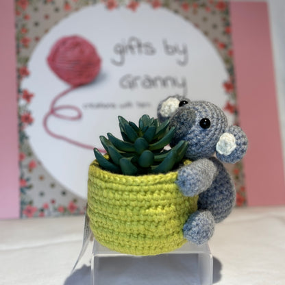 A handcrafted animal hugs plant pot holder, featuring a grey koala bear holding a moss green pot with a green cactus plant. The item is displayed on a white stand with a floral patterned backdrop.