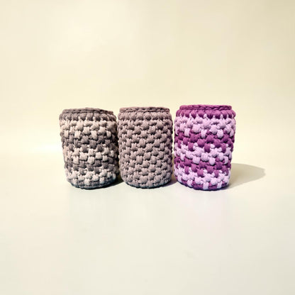 Three crocheted handmade storage pots on in silver and stone grey, one plain stone grey and finally one in purple and lilac, on a plain background