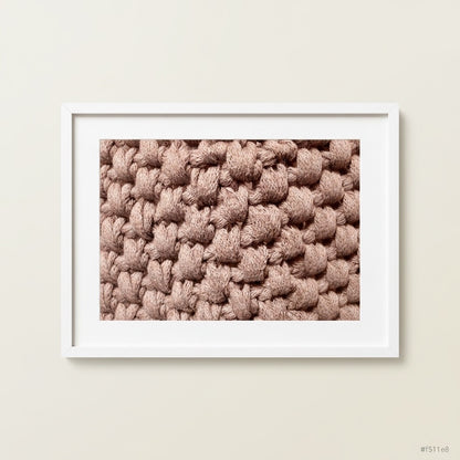 A framed art print showcasing a close-up of crochet stitches of a handmade storage pot in colour Ecru, set in a clean white frame against a neutral light beige wall.