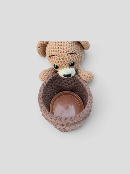 A top-down view of a brown crocheted pant pot cover with a beige crocheted teddy bear holding the plant pot cover. Inside the plant pot cover is a plastic saucer to hold a real plant. Set against a solid light grey background.