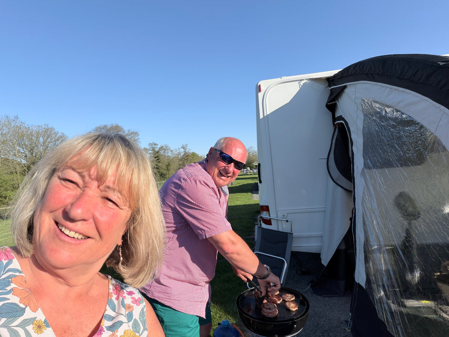 A smiling blonde woman takes a selfie next to a man grilling outdoors beside a white caravan under a clear blue sky.