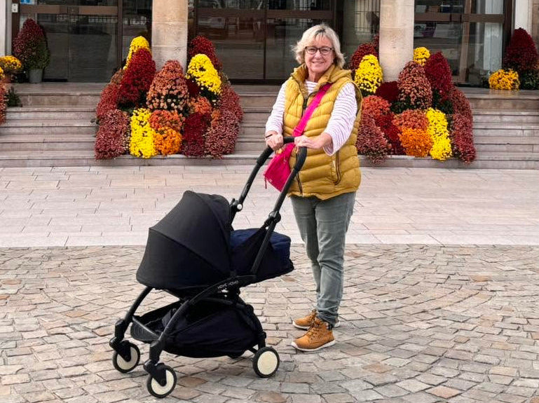 A woman stands in front of a historic building with floral decorations, pushing a black stroller. The building has stone columns and the words 'LIBERTE', 'EGALITE', and 'FRATERNITE' above the entrance.