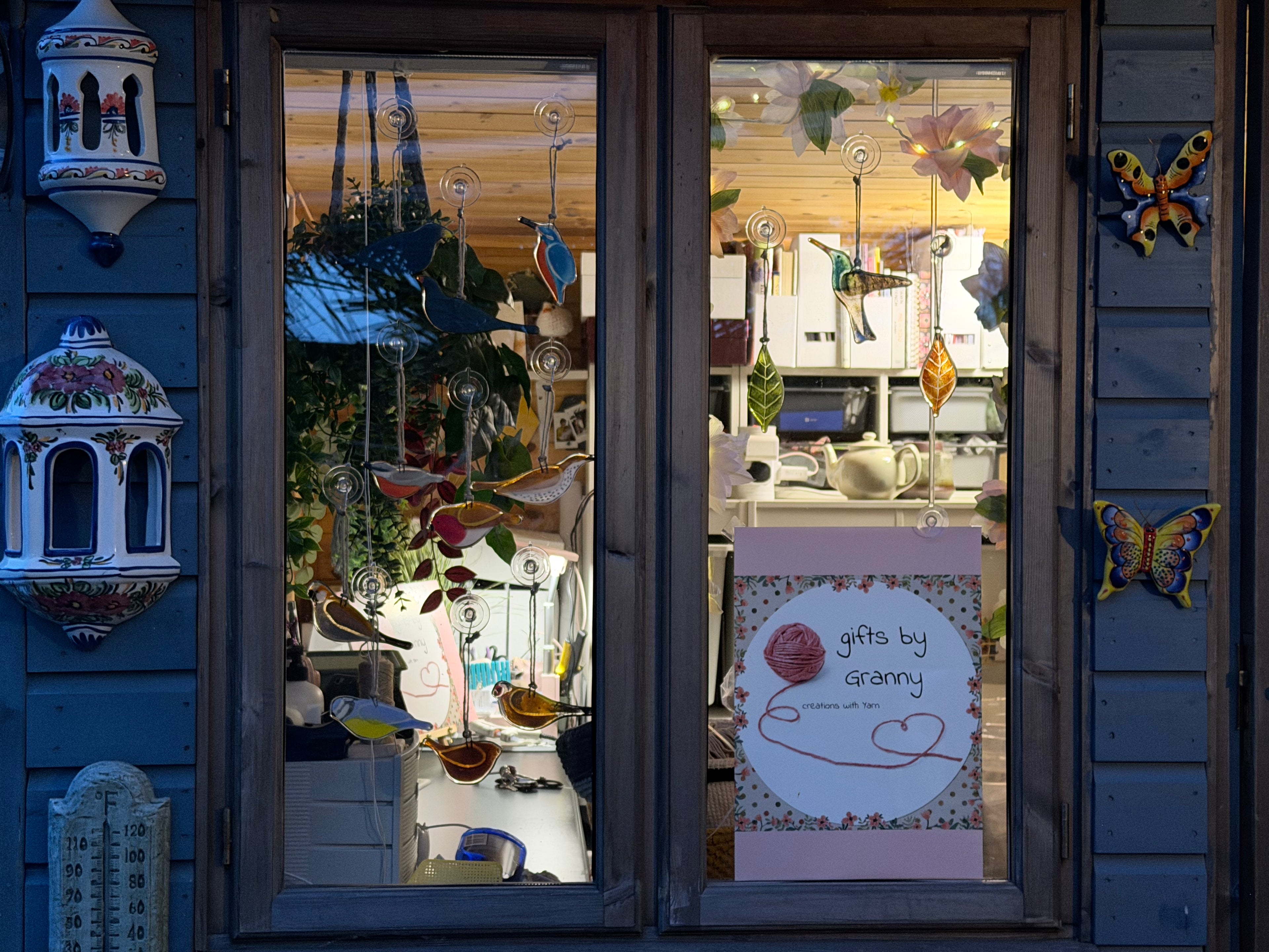 A storefront window display with blue wooden siding and hanging decorative items, including a butterfly ornament on the right, a bird ornament, and a sign reading 'gifts by granny.' The interior shows more handmade decorations and home goods behind glass.